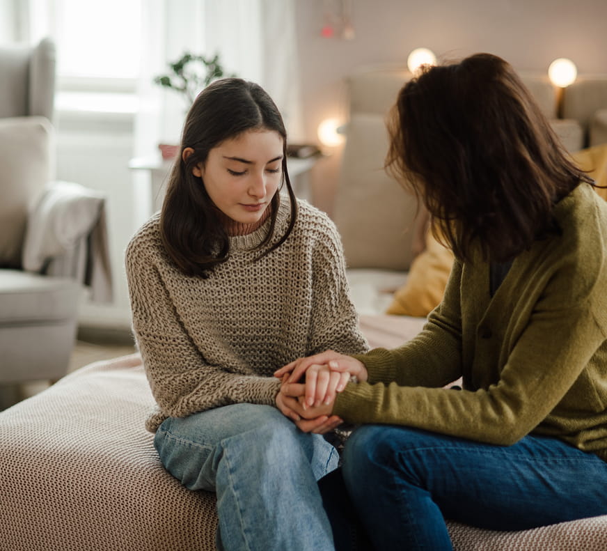 2 women on a bed, one is holding the others hands and is trying to comfort them.