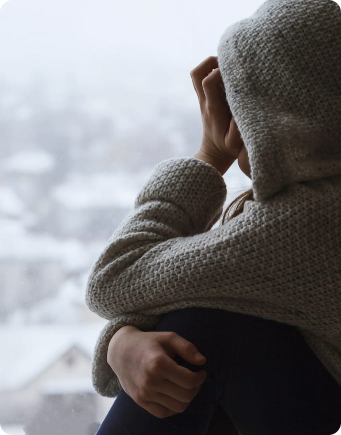 someone looking off into a snowy neighborhood as they are wearing a hoodie, holding their knees and are distressed about something