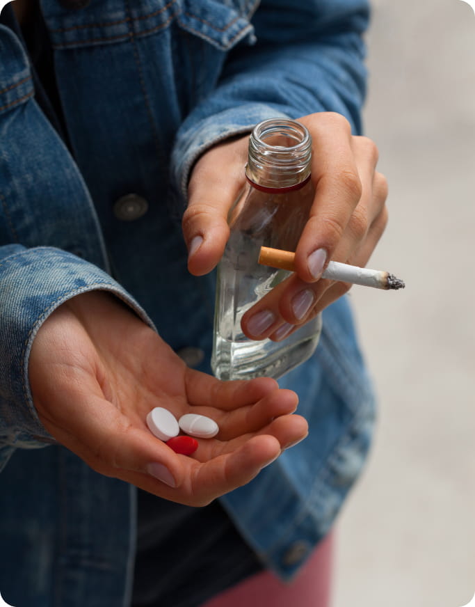 a closeup of someone holding pills, an alcohol bottle, as well as a lit cigarette.