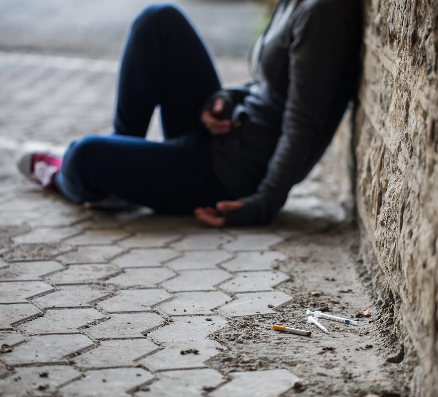 Someone sitting outside near an urban sidewalk with obvious drug needles and a cigarette nearby.