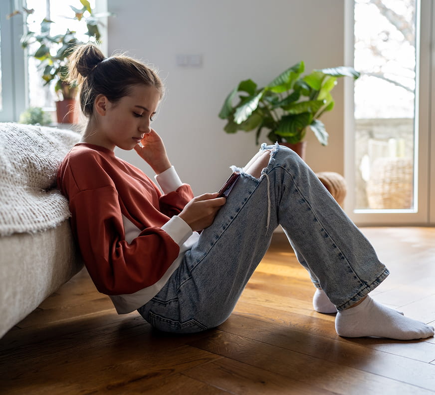 Support That Respects Your Teen - Girl sitting on the floor near the foot of her bed on her phone, looking distressed
