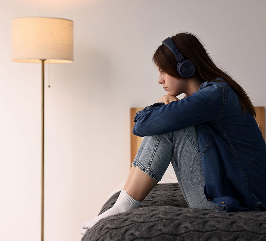 Trauma & PTSD Treatment - Girl sitting on her bed, arms hugging her knees and headphones are on, going through something stressful.