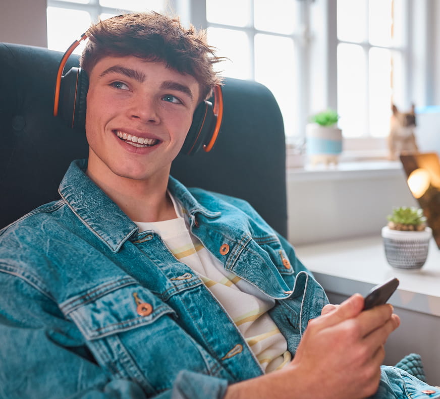 Boy sitting, smiling, with his phone up and in view with headphones on.